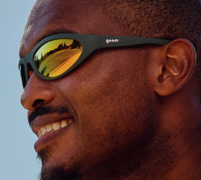 Smiling man wearing goodr Grass Stain Hall of Fame sunglasses with green wrap-around frame and amber mirrored lenses, perfect for running, cycling, and active outdoor sports.