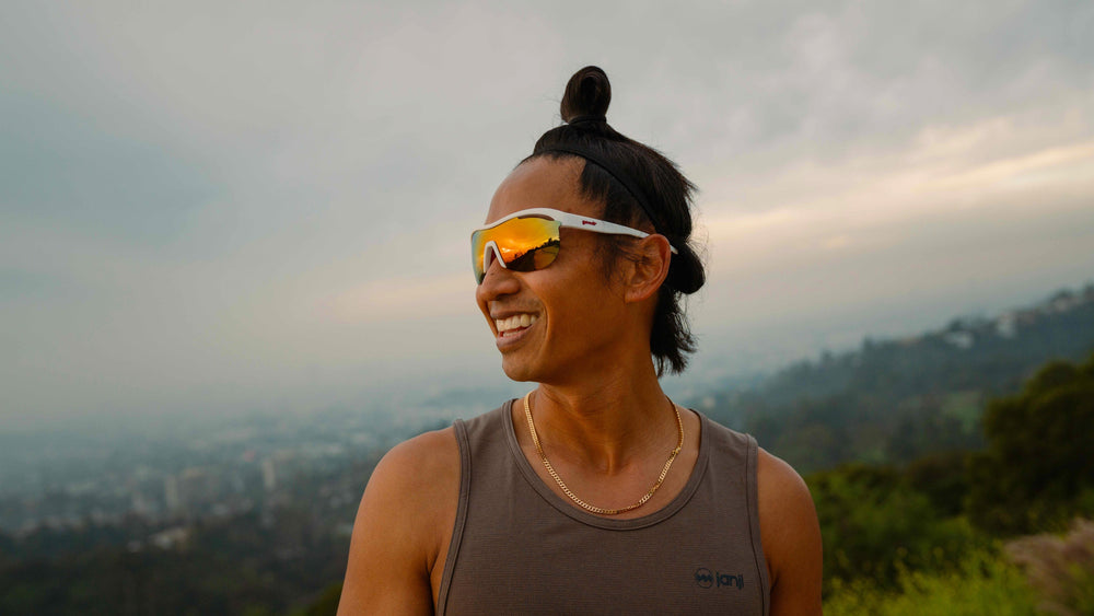 Smiling runner wearing Goodr Sonic G “Today’s Special: My Dust” sunglasses with mirrored lenses and white frame, standing outdoors at sunset with scenic mountain and city view, lightweight anti-slip performance eyewear for running and active lifestyle